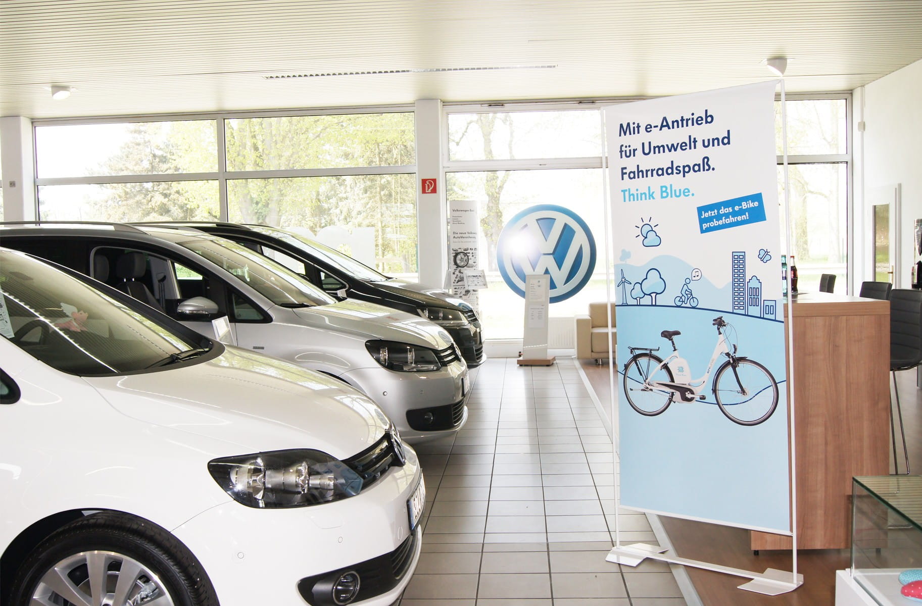 Roll-Up Easymag Stand von ENTDECKER mit dem Slogan 'Mit Antrieb für Umwelt und Fahrvergnügen. Think Blue.' im VW Autohaus, im Hintergrund mehrere VW Fahrzeuge.