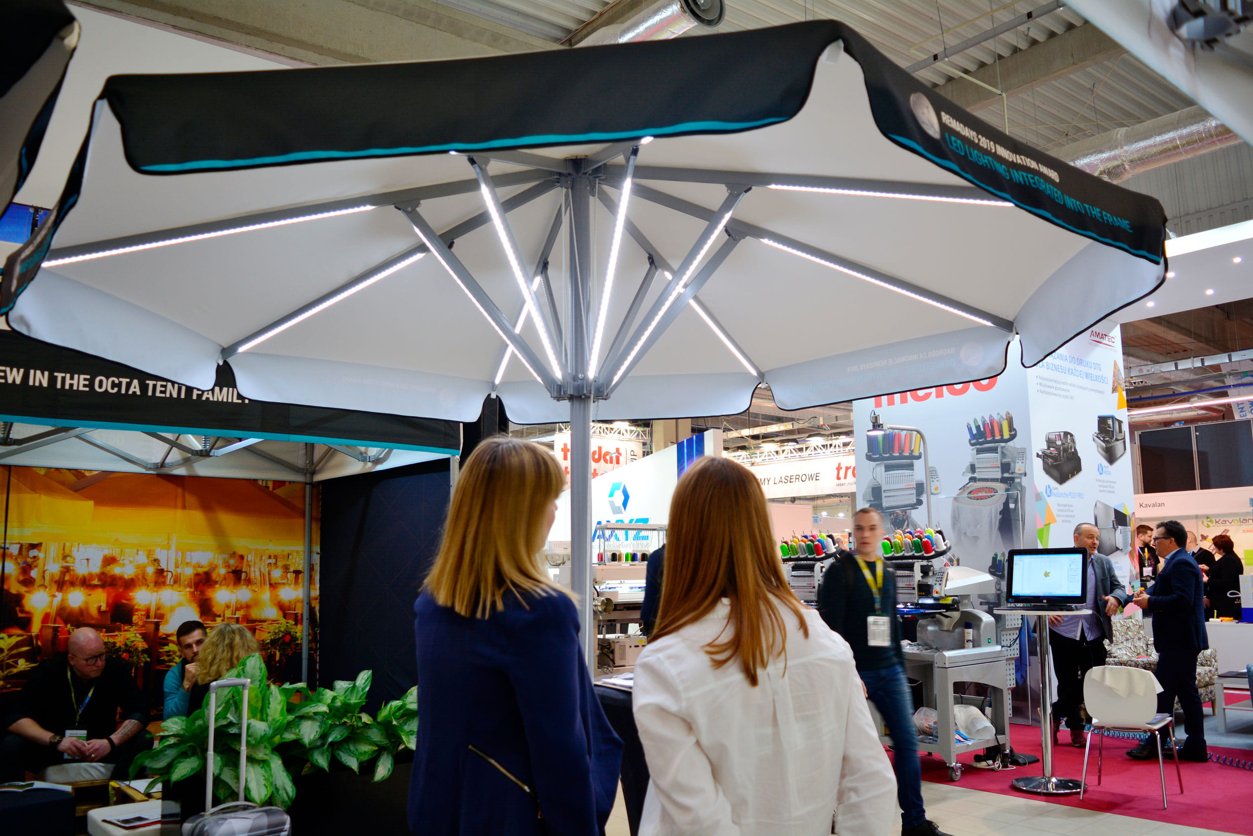 Zwei Personen betrachten den Sonnenschirm Expert Led Messe am Stand, umgeben von Ausstellungsständen.