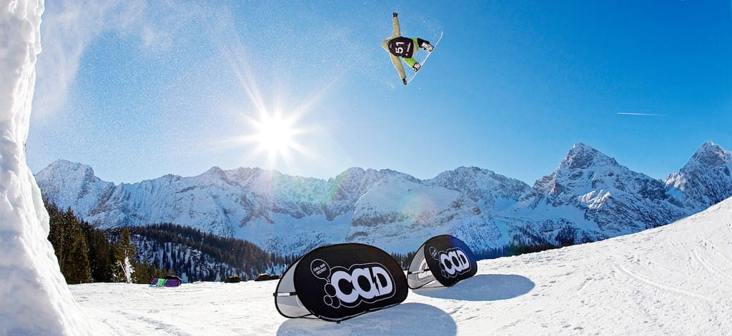 Snowboarder springt über eine Werbebande im Schnee mit Bergen im Hintergrund.
