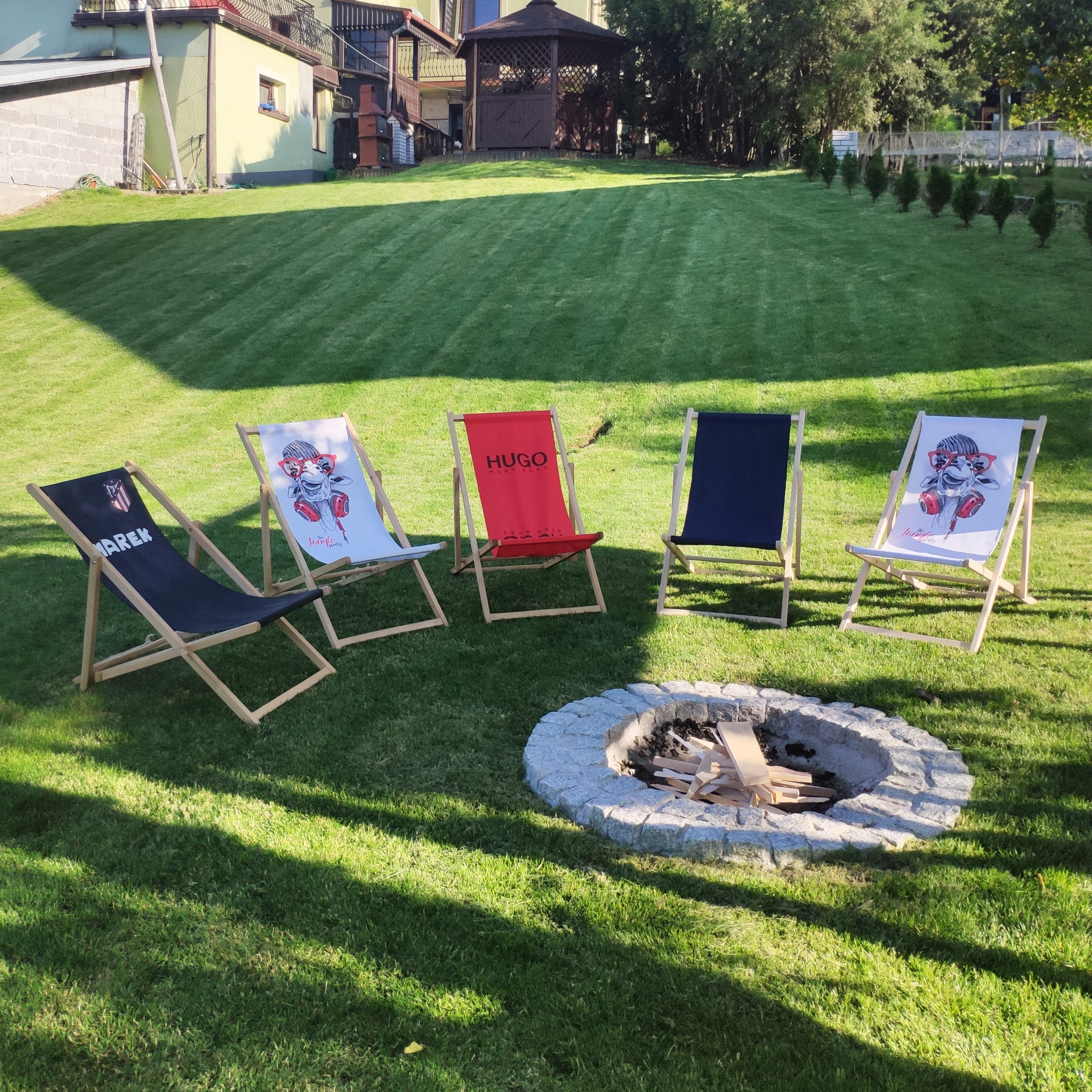 Fünf Liegestühle im Garten um einen Steinkreis mit Holz, zwei Stühle mit Aufdrucken von 'HUGO' und 'DREW', Rasenfläche im Hintergrund.