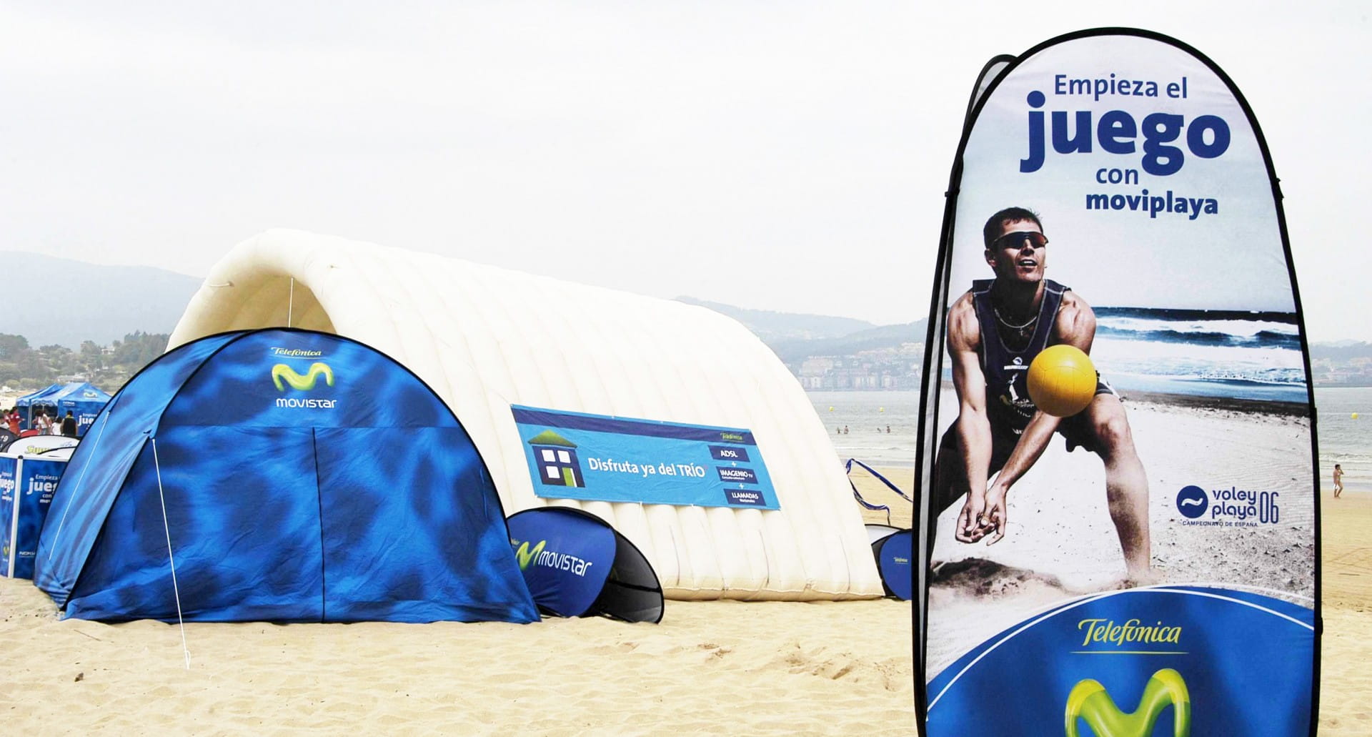 Werbesäule 'Quicktower' von Telefonica am Strand, mit einem Mann, der Beachvolleyball spielt. Im Hintergrund ein blauer Pavillon.