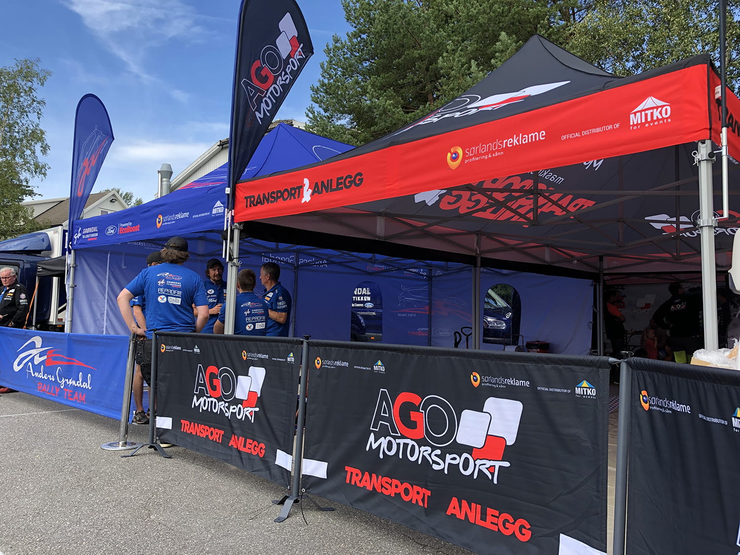 Zelt und Banner von AGO Motorsport bei einer Motorsportveranstaltung, mit Teammitgliedern in blauen Shirts.