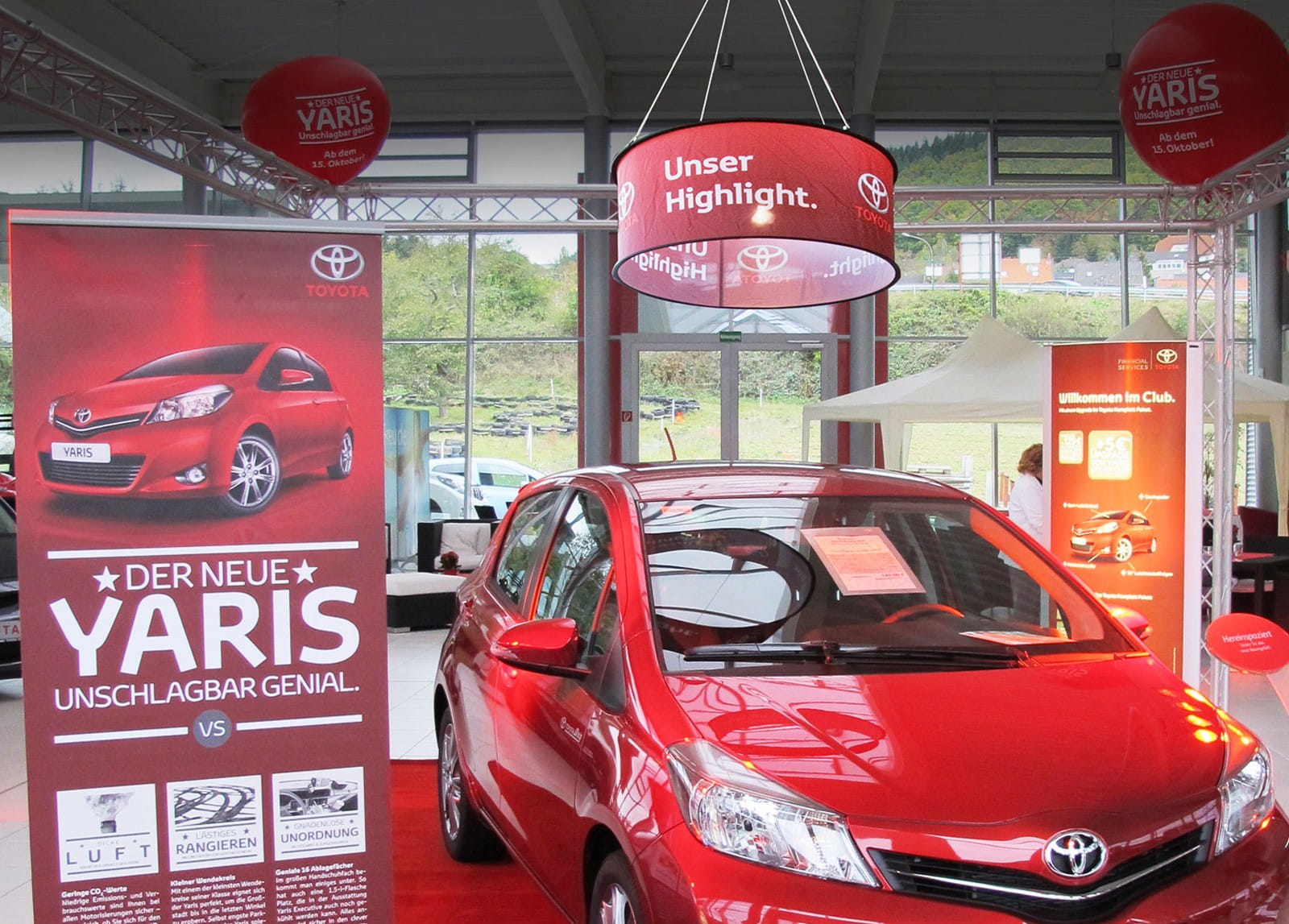 Deckenhänger mit dem Text 'Unser Highlight' und Toyota-Logo, über einem roten Toyota Yaris in einer Ausstellungsumgebung.