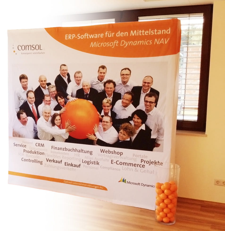 Messewand mit dem Produktnamen 'Quickup Stoffwand Comsol', zeigt ein Team von Personen vor einem großen orangefarbenen Ball, mit Text über ERP-Software für den Mittelstand und Microsoft Dynamics NAV.