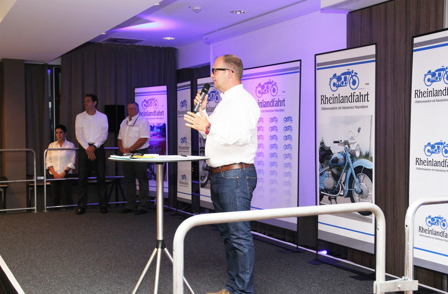 Mann spricht am Mikrofon vor Roll-Up Bannern des Produkts 'Einsatzbeispiel Roll Up Easymag Stand Rheinlandfahr'.