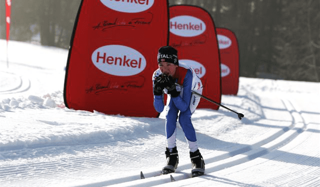 Skilangläufer in blauer und weißer Sportkleidung, der auf einer schneebedeckten Strecke fährt, mit Werbesäulen von Henkel im Hintergrund.