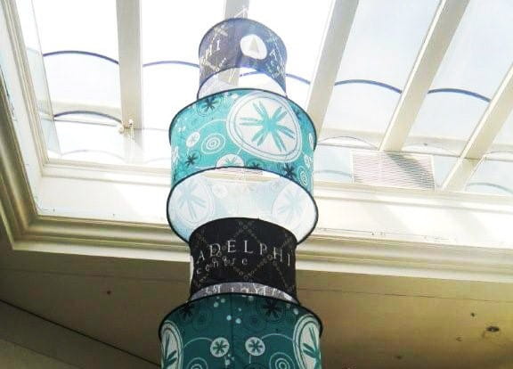 Deckenhänger Cylindrical Flexibel von ENTDECKER, bestehend aus mehreren übereinander gestapelten zylindrischen Elementen in verschiedenen Farben und Mustern, aufgehängt unter einem Glasdach.