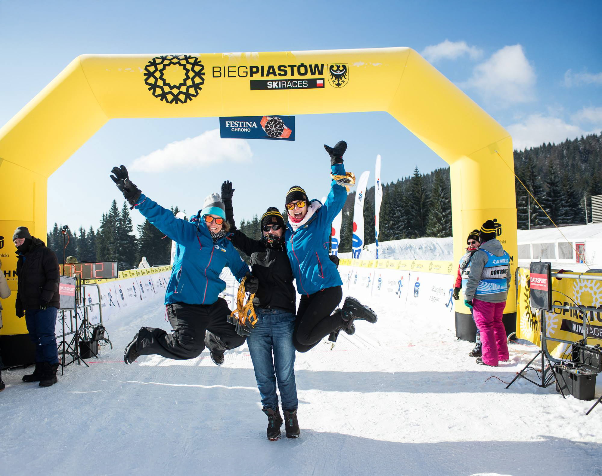 Drei Personen feiern unter einem gelben Torbogen mit der Aufschrift 'BIEGPIASTOW SKIRACES' im Schnee.