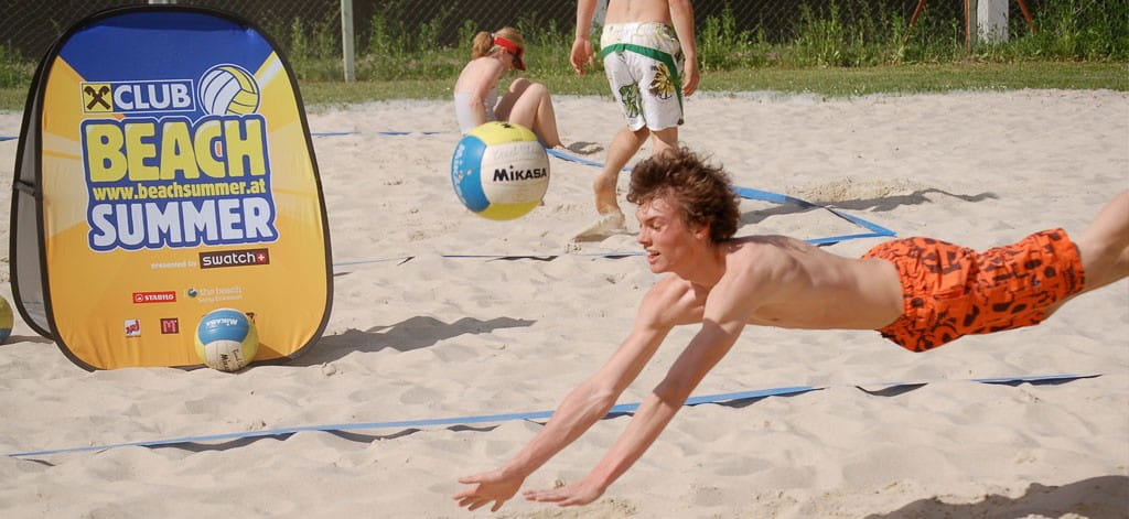 Ein Junge mit orangen Shorts springt im Sand, um einen Volleyball zu erreichen. Im Hintergrund sind weitere Spieler und eine Werbebande für 'BEACH SUMMER' zu sehen.