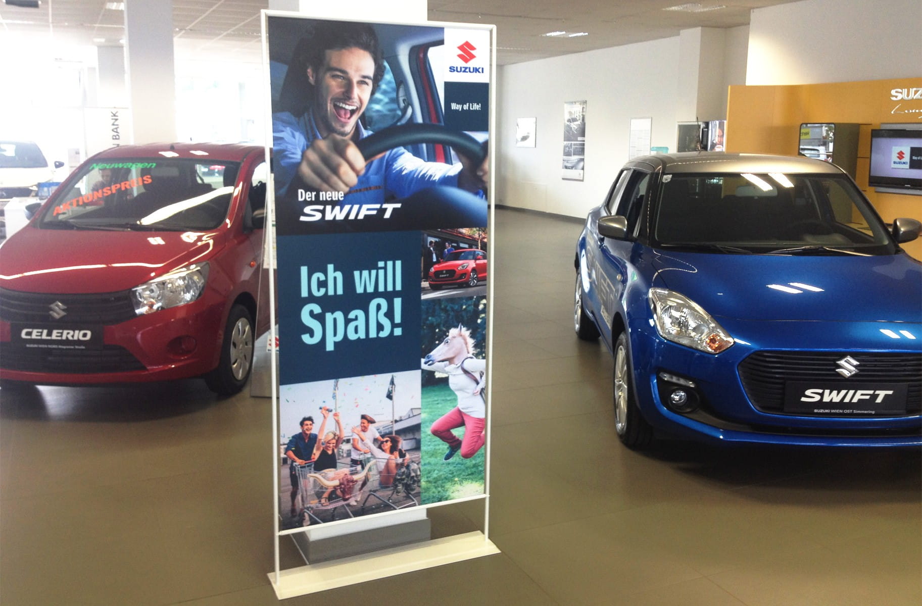 Roll-Up Easymag Stand von Suzuki mit dem Slogan 'Ich will Spaß!' und Bildern von glücklichen Menschen, platziert zwischen einem roten und einem blauen Suzuki Swift.