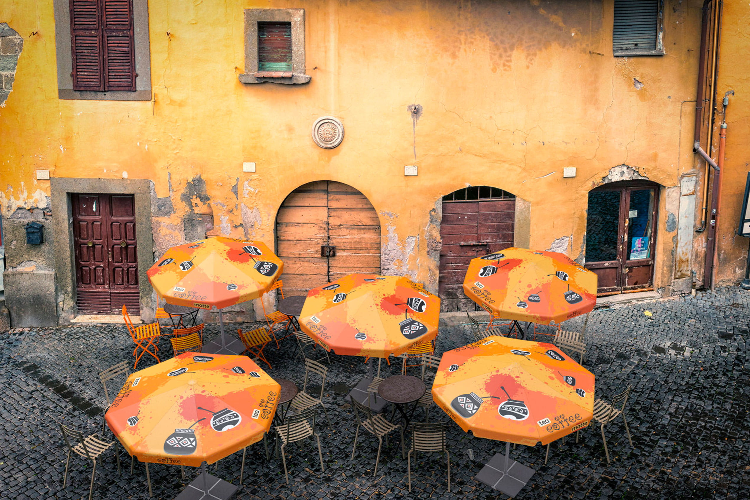 Sonnenschirm Expert Cafe in orange, auf einem gepflasterten Platz vor einer gelben Wand mit alten Fenstern.