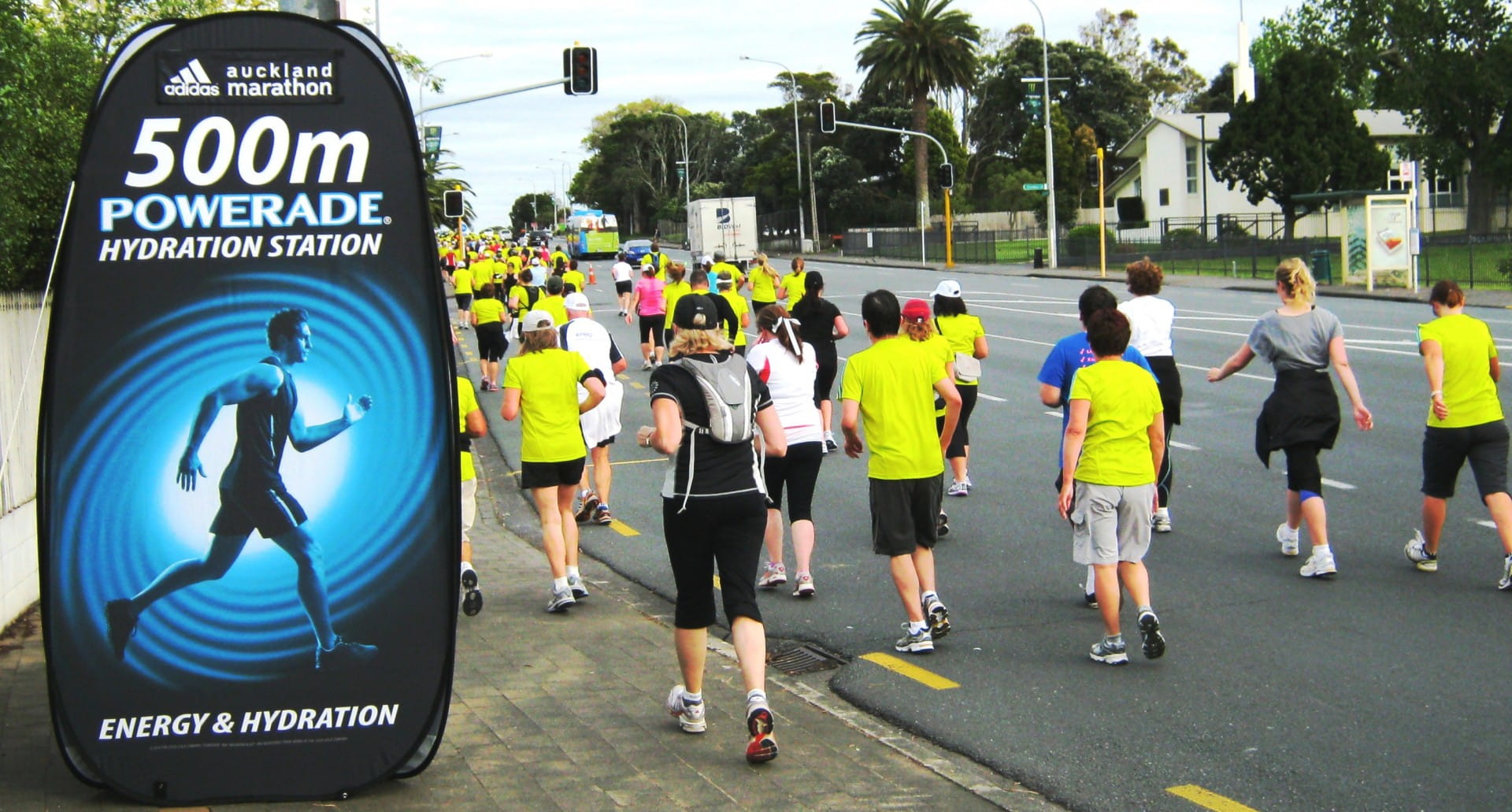 Werbesäule für Powerade bei einem Marathon, zeigt '500m POWERADE HYDRATION STATION' und Läufer in gelben Shirts.