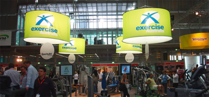 Beleuchtete zylindrische Deckenhänger mit dem Schriftzug 'exercise Australia' über einem Fitness-Messebereich.