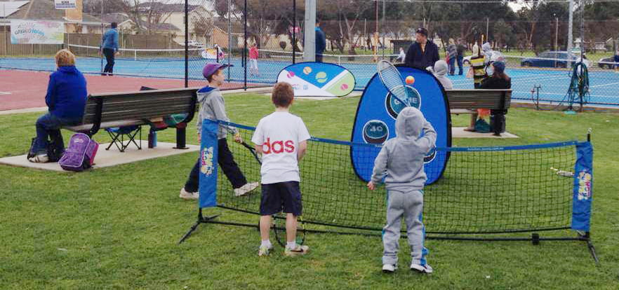 Zwei Kinder spielen mit einem Tennisball an einem Quickboard Werbebande auf einem Tennisplatz, während Erwachsene im Hintergrund zuschauen.