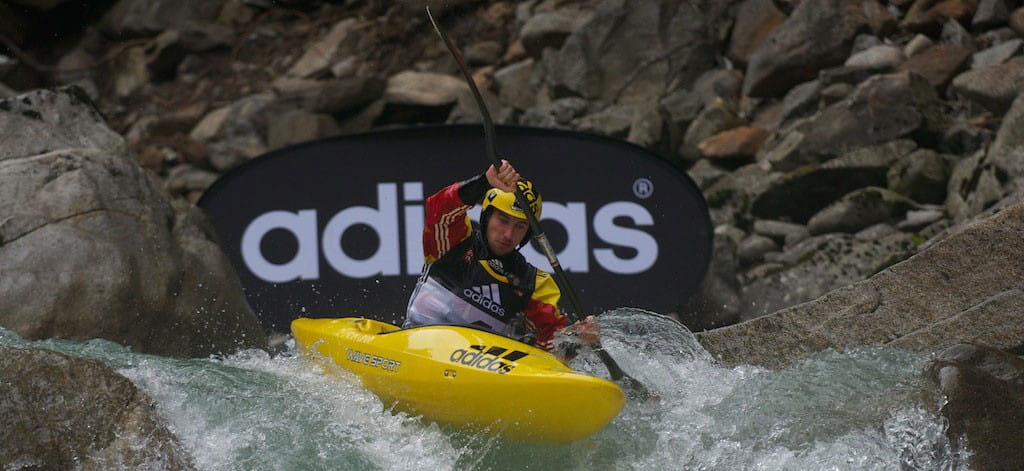Ein Kanufahrer in einem gelben Kanu paddelt durch eine reißende Strömung, im Hintergrund ist ein Werbebanner von adidas sichtbar.