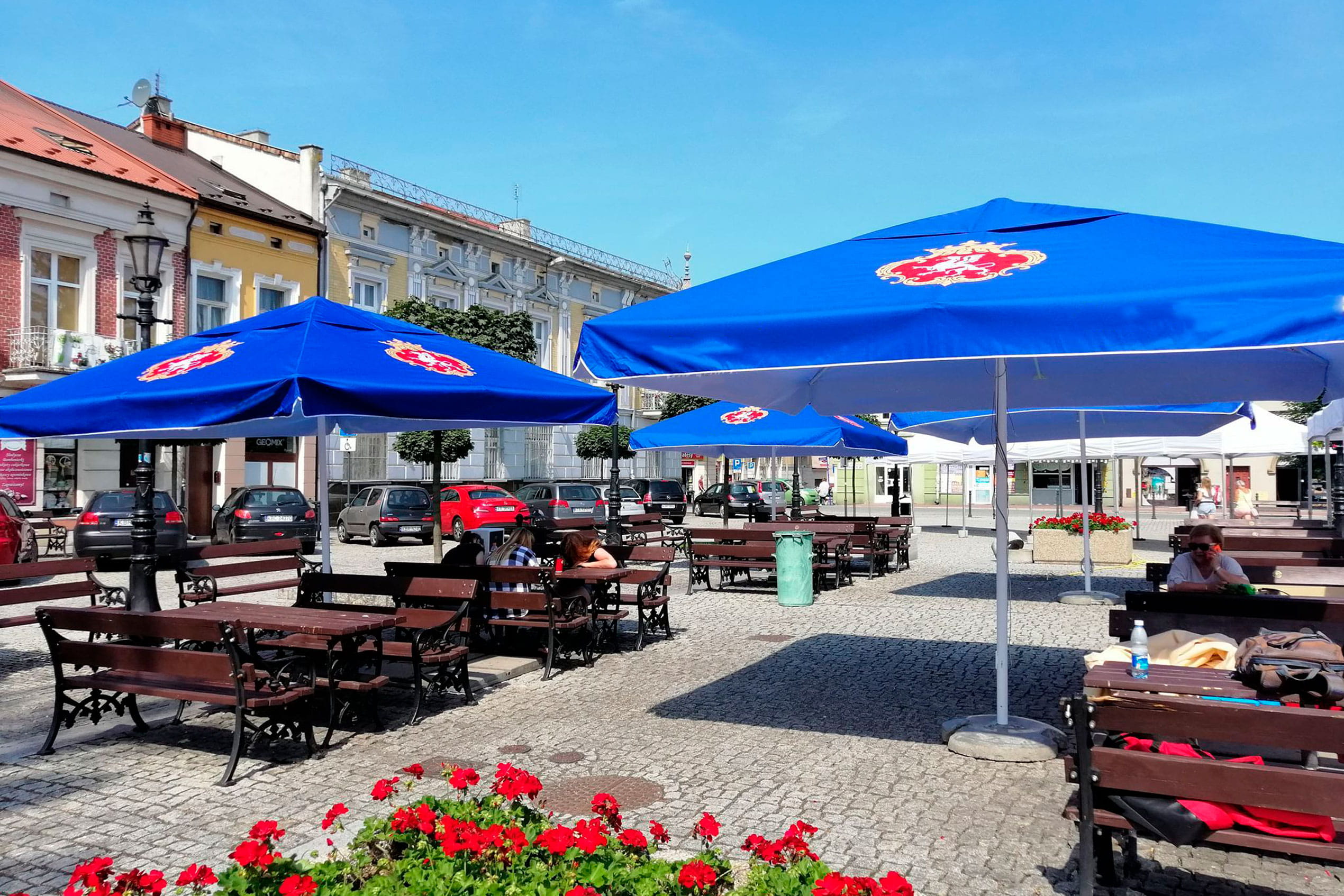Biergarten mit blauen Sonnenschirmen des Produkts 'Sonnenschirm Expert Biergarten', Tischen und Stühlen auf einem Platz.